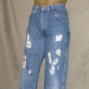 May include: Light blue denim jeans with distressed detailing. The jeans have multiple rips and tears across the front of the legs, giving them a worn and vintage look. The jeans have a classic five-pocket design and a button and zip closure.