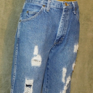 May include: Light blue denim jeans with distressed detailing. The jeans have multiple rips and tears across the front of the legs. They feature a classic five-pocket design and a button and zip closure.