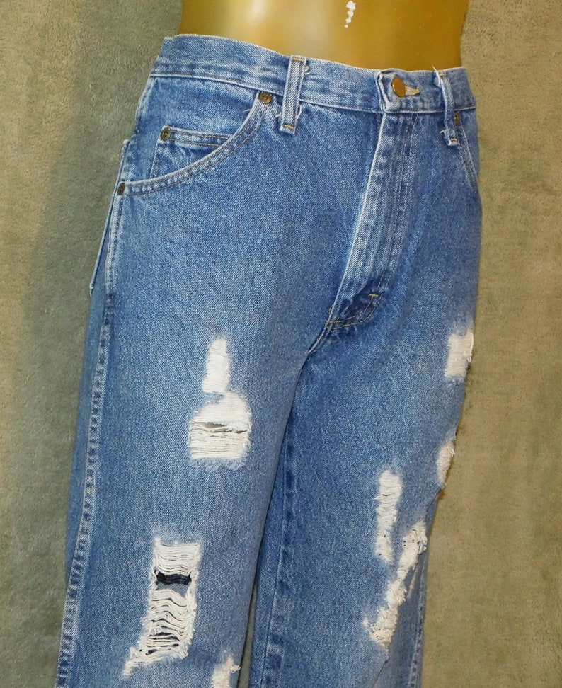 May include: Light blue denim jeans with distressed detailing. The jeans have multiple rips and tears across the front of the legs. They feature a classic five-pocket design and a button and zip closure.