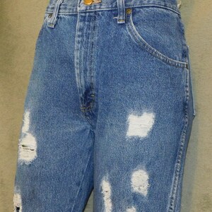 May include: Blue denim jeans with distressed detailing. The jeans feature multiple rips and tears across the front of the legs, creating a worn and vintage aesthetic. The jeans have a classic five-pocket design and a button and zip closure.