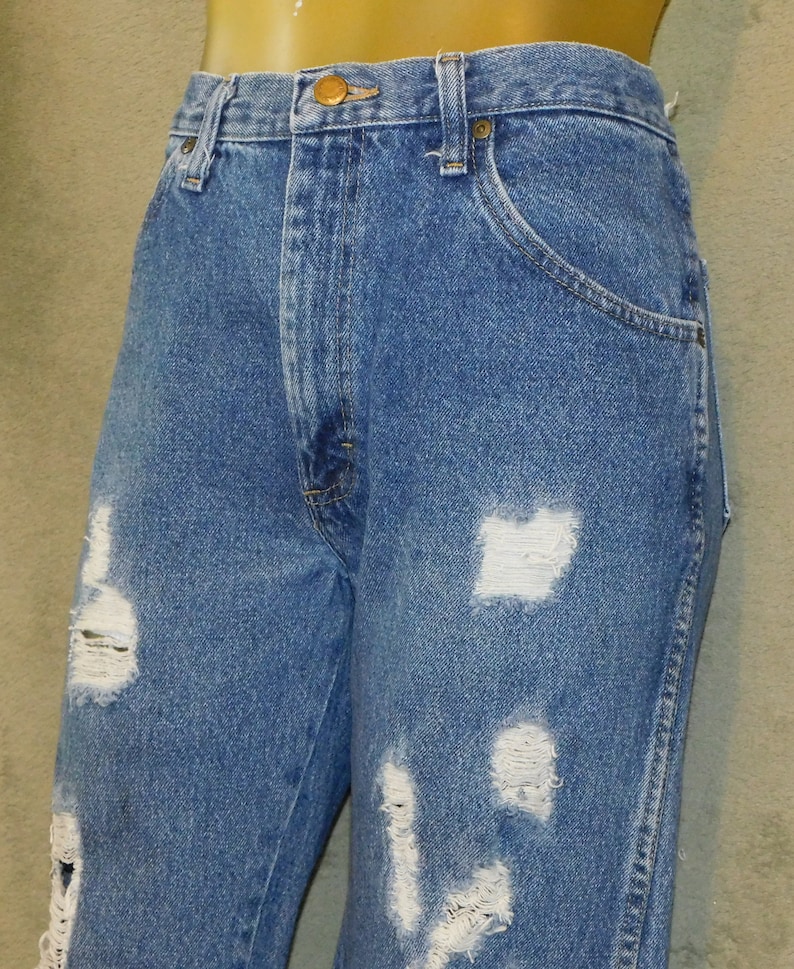 May include: Blue denim jeans with distressed detailing. The jeans feature multiple rips and tears across the front of the legs, creating a worn and vintage aesthetic. The jeans have a classic five-pocket design and a button and zip closure.