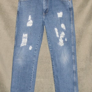 May include: Light blue denim jeans with distressed detailing. The jeans have multiple ripped areas across the front of the legs. The jeans are hanging on a metal hanger. The jeans have a classic five-pocket design.