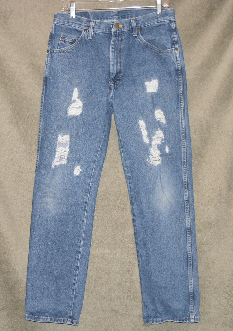 May include: Light blue denim jeans with distressed detailing. The jeans have multiple ripped areas across the front of the legs. The jeans are hanging on a metal hanger. The jeans have a classic five-pocket design.