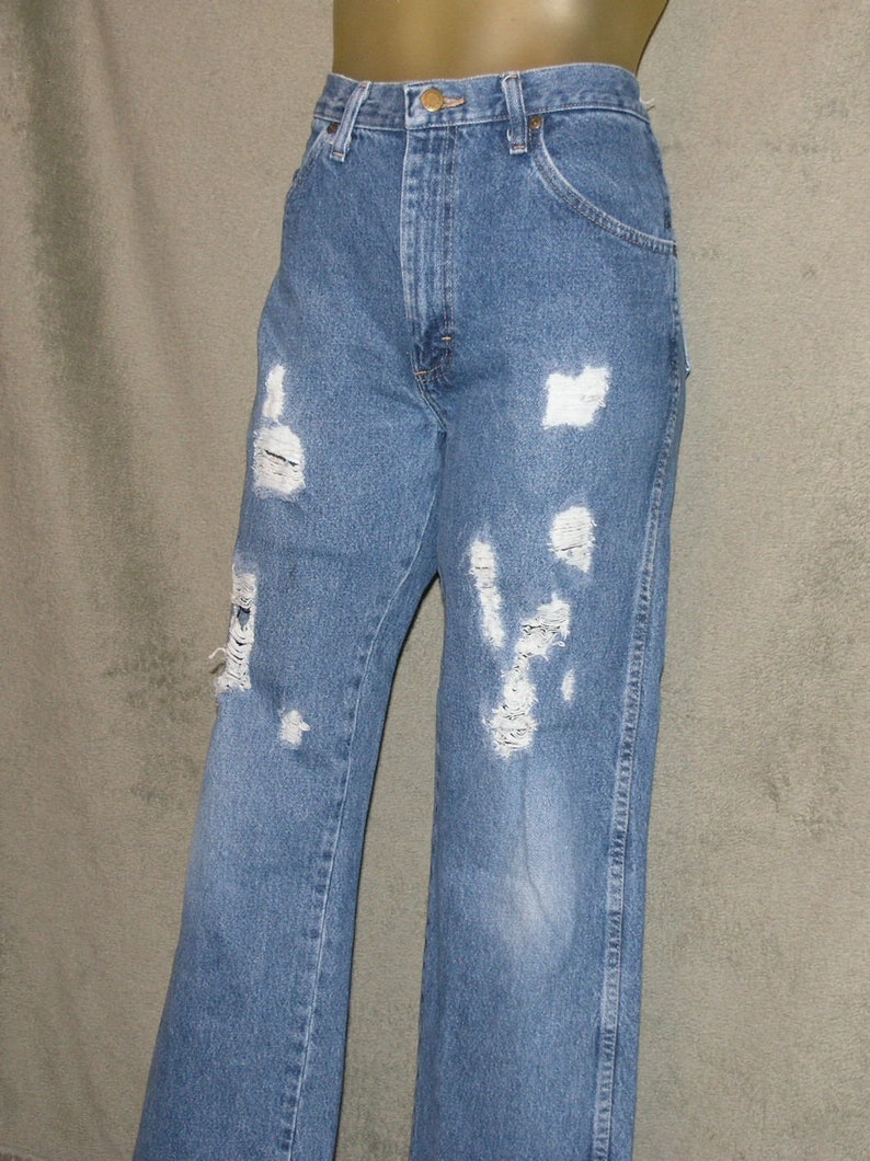 May include: Light blue denim jeans with distressed detailing. The jeans have multiple rips and tears across the front of the legs, giving them a worn and vintage look. The jeans have a classic five-pocket design and a button and zip closure.