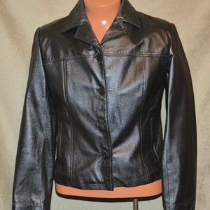 May include: A black leather jacket featuring a classic collar, long sleeves, and a front button closure with three visible black buttons. The jacket has a structured fit with vertical seam detailing on the front panels and a smooth, polished finish.