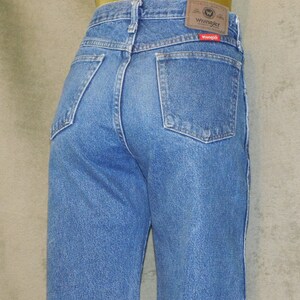 May include: Light blue denim jeans with a classic five-pocket design. The jeans have a straight leg cut and a Wrangler brand patch on the back waistband. The back pockets are a standard square shape.