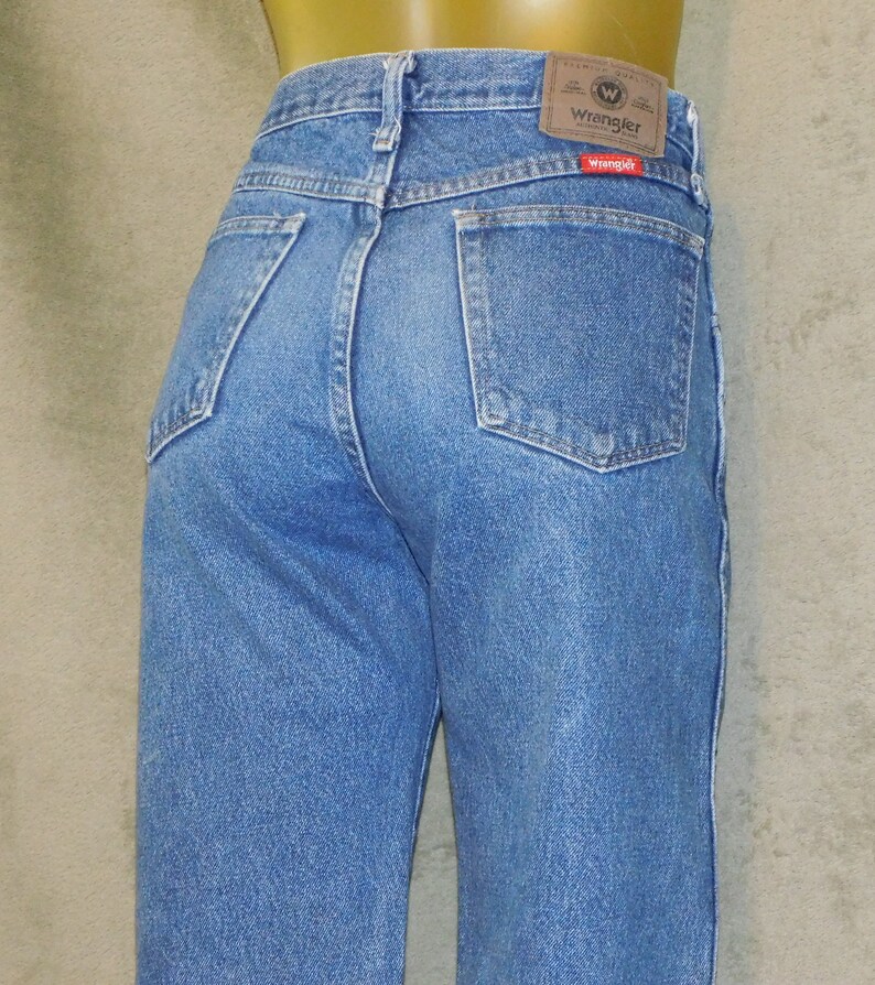 May include: Light blue denim jeans with a classic five-pocket design. The jeans have a straight leg cut and a Wrangler brand patch on the back waistband. The back pockets are a standard square shape.