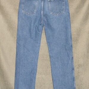 May include: Pale blue denim jeans with a classic five-pocket design. The back view shows the Wrangler brand patch and two back pockets. The jeans are hanging on a hanger.