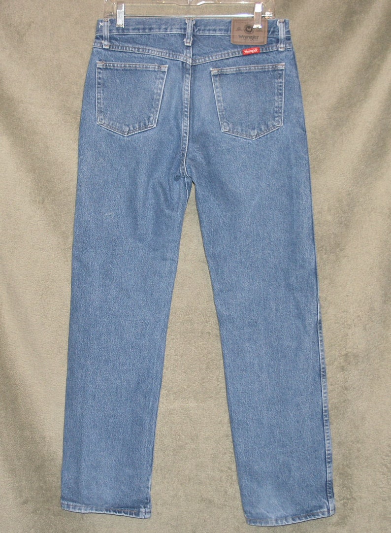 May include: Pale blue denim jeans with a classic five-pocket design. The back view shows the Wrangler brand patch and two back pockets. The jeans are hanging on a hanger.