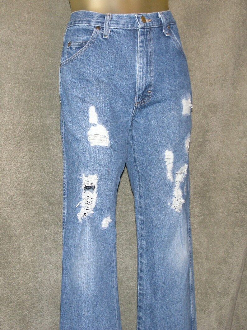 May include: Light blue denim jeans with distressed detailing. The jeans feature multiple ripped sections across the front of the legs, creating a worn and vintage aesthetic. The jeans have a classic five-pocket design.