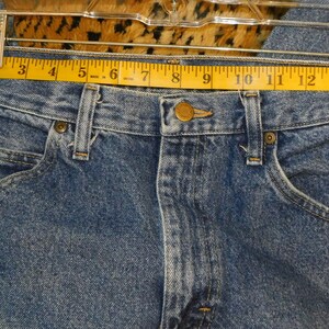 May include: Close-up of blue denim jeans with a yellow measuring tape across the waist. The jeans have a button closure, belt loops, and front pockets. The waist measures approximately 33 cm.