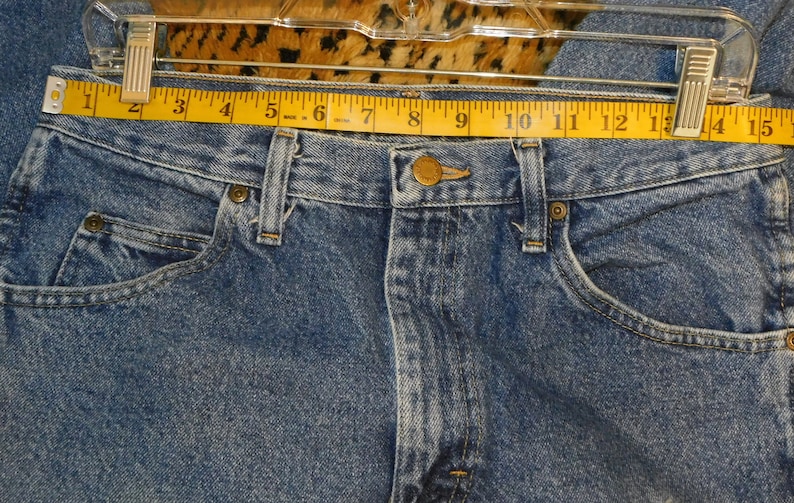May include: Close-up of blue denim jeans with a yellow measuring tape across the waist. The jeans have a button closure, belt loops, and front pockets. The waist measures approximately 33 cm.