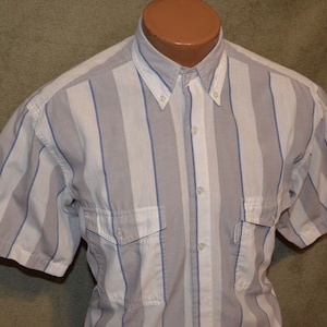 May include: Short-sleeved button-down shirt with vertical stripes in white, light gray, and blue. The shirt has two chest pockets and a button-down collar. The shirt is displayed on a mannequin.