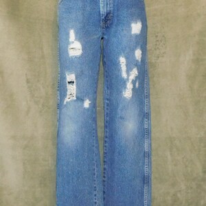 May include: Light blue denim jeans with distressed details. The jeans have a high waist and a wide leg. Several areas of the jeans are ripped and torn, with white patches. The jeans are displayed on a mannequin.