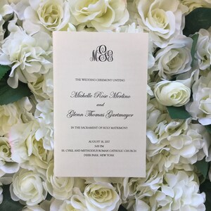 Puede incluir: Un programa de ceremonia de boda de color crema con escritura negra, con los nombres de Michelle Rose Merlino y Glenn Thomas Gartmayer. El programa está sobre un fondo de rosas blancas. El programa incluye la fecha, la hora y el lugar.
