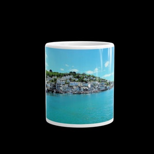 May include: A white ceramic mug with a colorful image of a seaside town with buildings and boats. The sky is blue with white clouds. The water is a bright turquoise color.