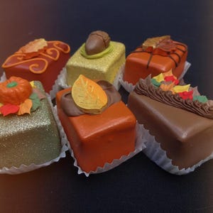 以下が含まれることがあります： 6つのミニチュア装飾ケーキの詰め合わせ。各ケーキは長方形で、カボチャ、葉、ドングリなど、秋をテーマにした装飾が施されています。ケーキはオレンジ、茶色、緑など様々な色で、小さな紙のドイリーの上に置かれています。