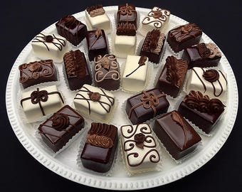 May include: A round white platter holds an assortment of rectangular petit fours. Each small cake is coated in either smooth dark chocolate or white icing, featuring decorative piped chocolate swirls, lines, and floral-shaped frosting accents.