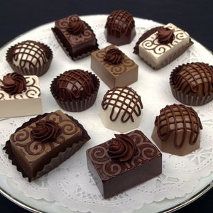 A white plate with a brown and white doily holds an assortment of chocolate fake candies and truffles. The candies are decorated with swirls and lines of chocolate.