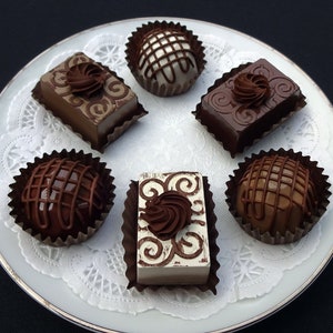A white plate with a lace doily holds six fake chocolate candies. The candies are in various shapes and sizes, including squares, circles, and ovals. Some of the candies are topped with swirls of chocolate.