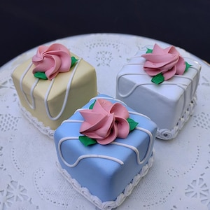 May include: Three miniature square cakes, each topped with a pink rose and green leaves. The cakes are in pastel colors: yellow, white, and blue, with white icing details and a decorative border. They sit on a white, lacy surface.