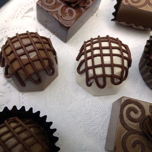 Assortment of gourmet chocolate faux candies with different designs and flavors. Some chocolates are topped with a lattice design, while others have swirls and a rosette design. The chocolates are arranged on a white surface with a lace pattern.