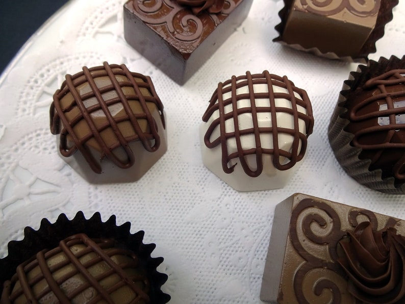 Assortment of gourmet chocolate faux candies with different designs and flavors. Some chocolates are topped with a lattice design, while others have swirls and a rosette design. The chocolates are arranged on a white surface with a lace pattern.
