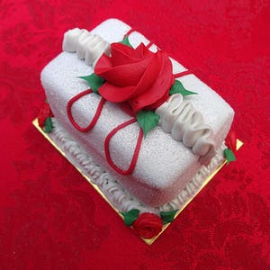 May include: A rectangular white cake covered in sparkling sugar, decorated with a large red rose, green leaves, and red ribbon accents. The cake sits on a gold-colored base and is trimmed with white frosting and small red roses.