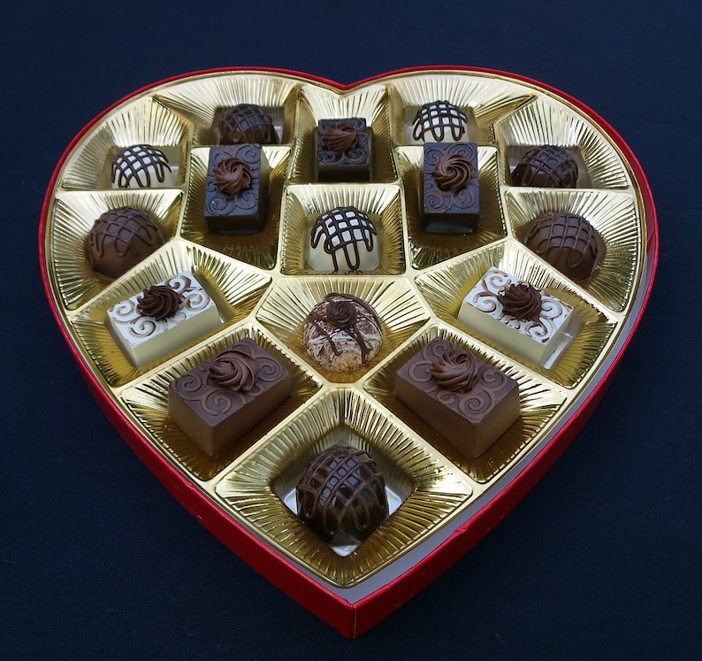 A heart-shaped box filled with assorted fake chocolates. The chocolates are arranged in a grid pattern and are a variety of colors and shapes.