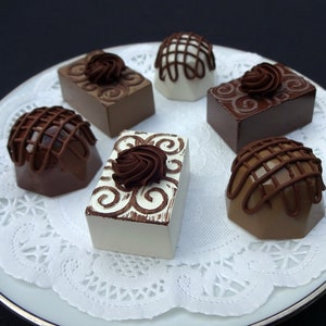 A white plate with a white doily on top. Six chocolate faux candies are arranged on the doily. The candies are different shapes and sizes, and some are decorated with swirls and lines.