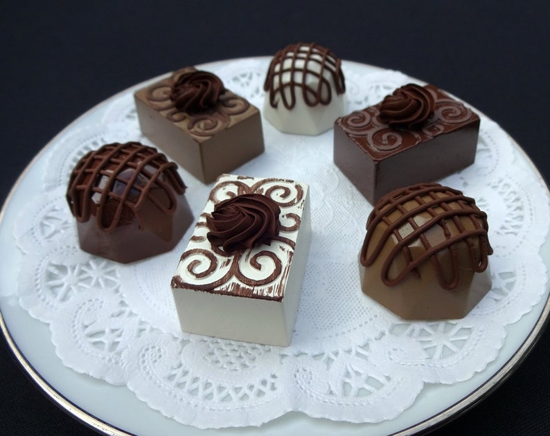 A white plate with a white doily on top. Six chocolate faux candies are arranged on the doily. The candies are different shapes and sizes, and some are decorated with swirls and lines.