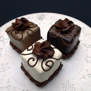 Op de afbeelding: Drie miniatuur chocoladetaarten versierd met swirls van chocolade glazuur en witte frosting. De taarten zijn gerangschikt op een witte kant.