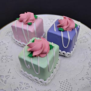 May include: Three square, pastel-colored cakes with pink frosting roses on top. The cakes are light pink, lavender, and mint green, with white icing stripes and decorative borders. They sit on a white, lacy doily.