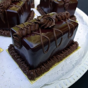 Könnte beinhalten: Nahaufnahme von mehreren rechteckigen Schokoladenkuchen mit glänzender, dunkelbrauner Glasur. Jeder Kuchen ist mit aufgespritzter Schokoladencreme in einem Wirbel- und Linienmuster verziert. Die Kuchen liegen auf einem weißen Teller.