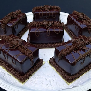 May include: Six rectangular chocolate pastries with glossy dark brown icing and decorative chocolate frosting. The pastries sit on individual gold squares atop a white, lacy paper doily on a white platter. A dessert.