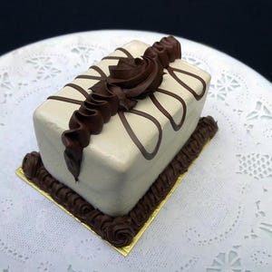 May include: A rectangular cake with a light beige frosting, decorated with dark brown chocolate swirls and a decorative chocolate ribbon. The cake sits on a gold-colored square base with chocolate frosting around the edges.