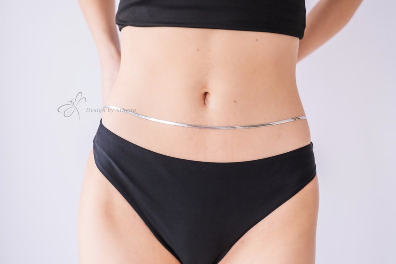 May include: A silver body chain worn around the waist. The chain is a thin, flat design. The model is wearing a black top and black underwear. The image has the text "Design by Athena" in the upper left corner.