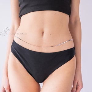May include: A silver body chain worn over black underwear. The chain is a delicate, thin design. The image also includes a black crop top. The text "Design by Athena" is visible in the upper left corner.