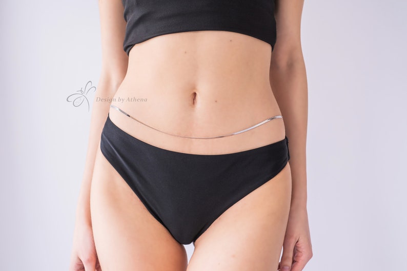 May include: A silver body chain worn over black underwear. The chain is a delicate, thin design. The image also includes a black crop top. The text "Design by Athena" is visible in the upper left corner.