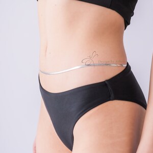 May include: A silver body chain with a flat, snake-like design. The chain is worn around the waist, resting on black underwear. The image includes the text "Design by Athenia".