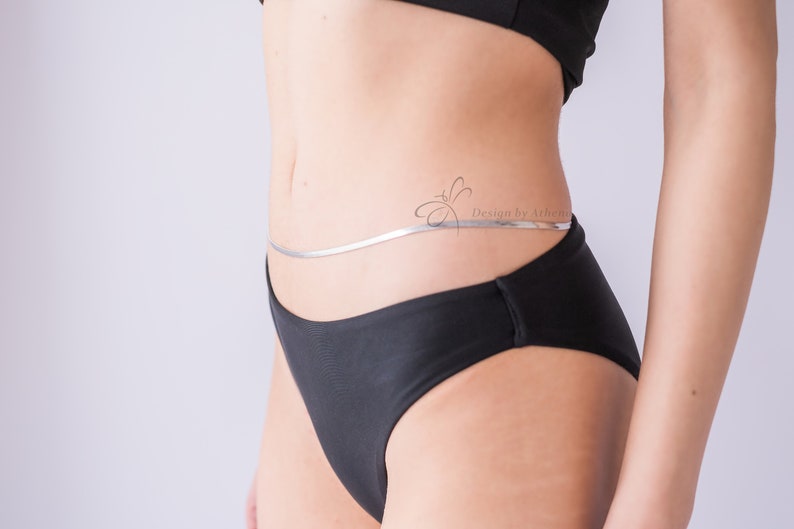 May include: A silver body chain with a flat, snake-like design. The chain is worn around the waist, resting on black underwear. The image includes the text "Design by Athenia".
