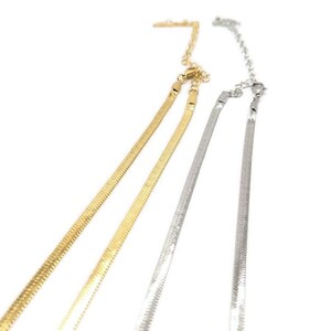 May include: Two herringbone chain necklaces, one gold-toned and one silver-toned, are displayed against a white background. The chains have a flat, scale-like design and adjustable clasps. The necklaces are likely for fashion or jewelry.