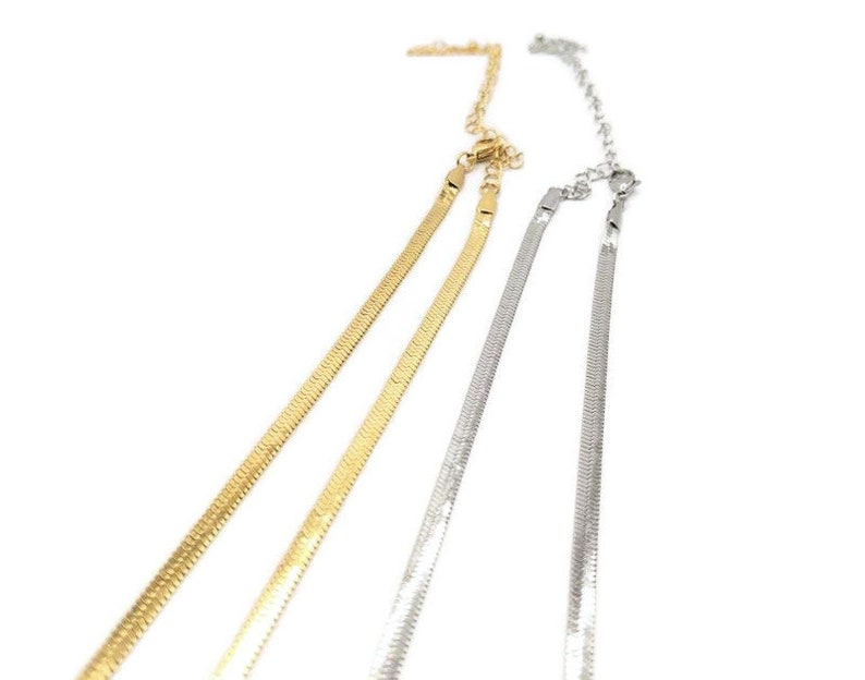 May include: Two herringbone chain necklaces, one gold-toned and one silver-toned, are displayed against a white background. The chains have a flat, scale-like design and adjustable clasps. The necklaces are likely for fashion or jewelry.