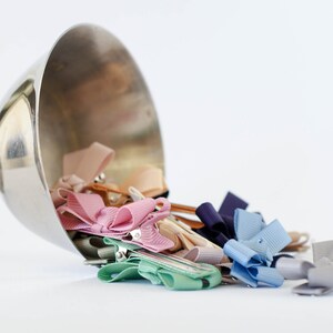 Op de afbeelding: Een zilveren beker die is omgevallen en een verzameling kleurrijke lint haarclips uitstort. De clips zijn in verschillende pastelkleuren, waaronder roze, blauw en beige. De achtergrond is effen wit.