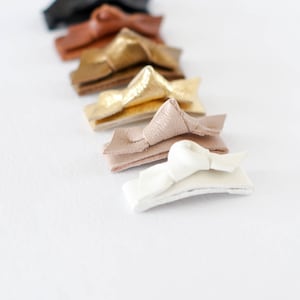 May include: A set of eight leather hair clips in various colors, including black, brown, gold, and white. The clips are arranged in a row, with the white clip at the bottom.