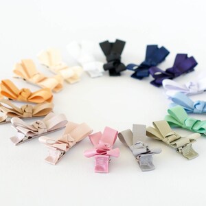 Op de afbeelding: Een verzameling kleine, kleurrijke strikhaarclips, gerangschikt in een cirkel. De strikken zijn in verschillende tinten, waaronder roze, blauw, groen en neutrale tinten. Elke clip heeft een metalen sluiting. De achtergrond is effen wit.