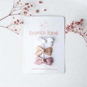 May include: Three small hair bows in white, tan, and mauve are displayed on a white card with the text "bambi lane hair accessories". The card is decorated with a logo and the website address www.bambilaneaccessories.etsy.com.