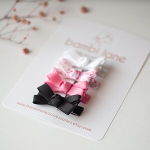 May include: A set of four small hair bows in black, pink, and floral patterns. The bows are arranged on a white card with the text "bambi lane accessories.etsy.com". The bows are made of ribbon and are likely for children.