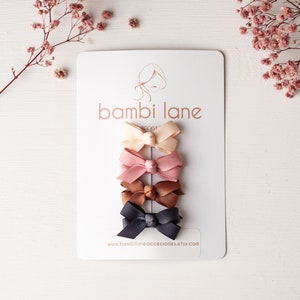 May include: Set of five mini hair bows in various colours, including beige, pink, brown, and black. The bows are arranged vertically on a white card with the text "bambi lane" and a website address.