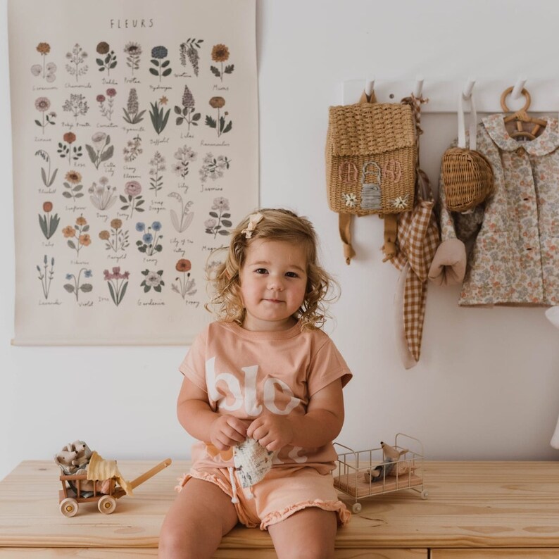 May include: A young child wearing a peach-colored t-shirt and shorts with the word "blo" printed on the shirt. A floral print poster hangs on the wall behind the child. A small wooden wagon and a metal toy cart are on the wooden surface.